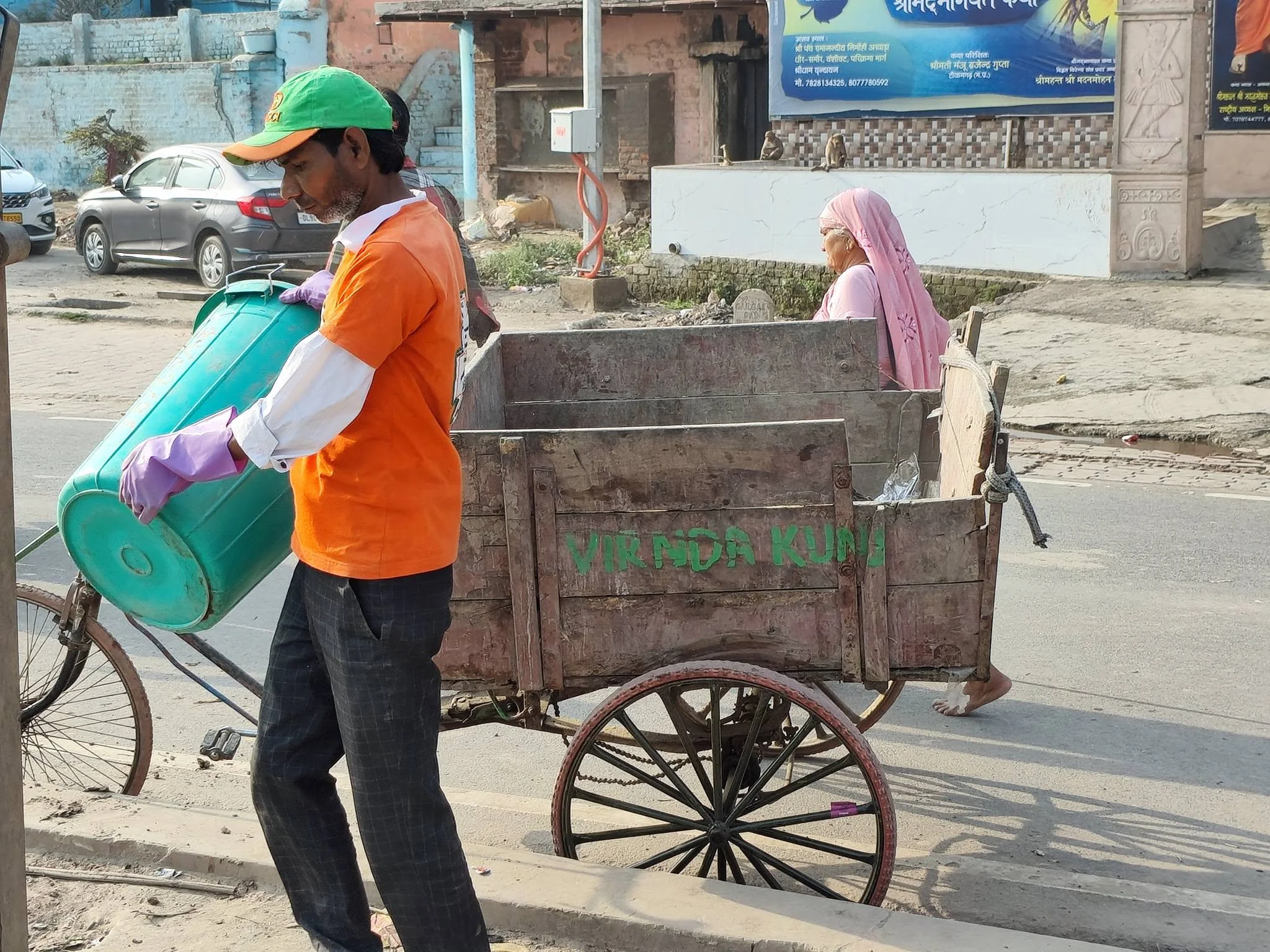 A worker in orange clothing handling a green waste container beside a collection cart