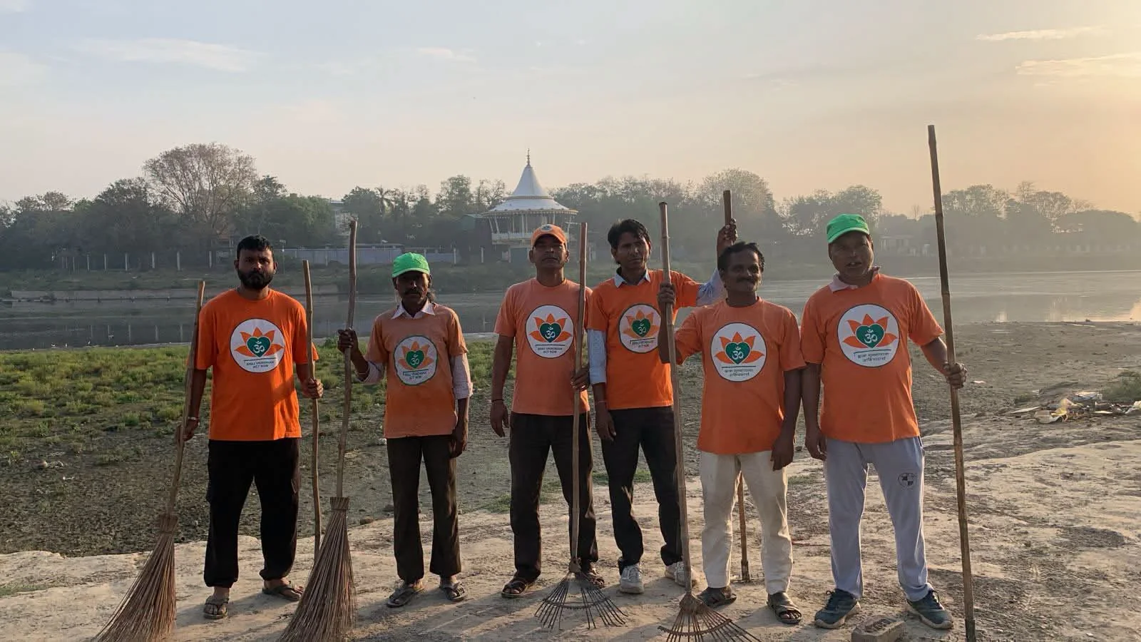 Five team members wearing orange project shirts and holding cleaning brooms beside a body of water